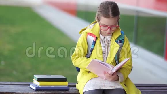 戴眼镜的冷静女孩坐在学校附近的长凳上学习功课这孩子穿着一件黄色夹克女生视频的预览图