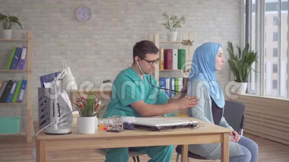 年轻的男医生用听诊器听一个戴头巾的年轻穆斯林妇女讲话视频的预览图