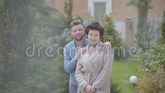 画像上幸福成熟的女人站在花园里的大房子前面成年的孙子抱着她把她放在视频的预览图