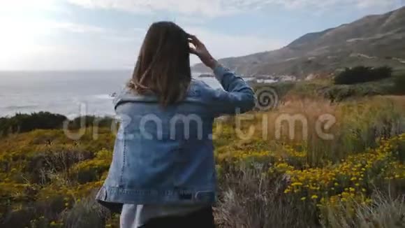 镜头跟随年轻快乐的女人走在黄色的花丛上走向大苏尔海洋海岸线上令人惊叹的岩石视频的预览图