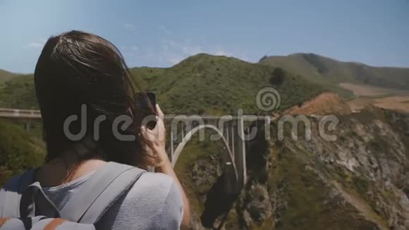 后景年轻的旅行者女士徒步旅行与背包在著名的比克斯比大桥拍摄史诗般风景的智能手机照片视频的预览图