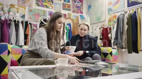 年轻女子向喝咖啡的成年女子展示杂志视频的预览图