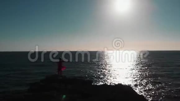 女人的轮廓站在悬崖上双手分开在空中对抗太阳和蓝色海洋上的太阳之路有手臂的女孩的无人视角视频的预览图