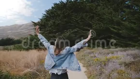 镜头跟随美丽的年轻快乐的自由女人奔跑手臂张开在一条小路上进入大苏尔黑暗的森林视频的预览图