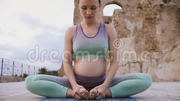 年轻的孕妇在户外做瑜伽坐着莲花姿势呼吸练习视频的预览图