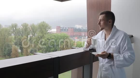 一个穿浴袍的年轻人正在阳台上喝咖啡视频的预览图