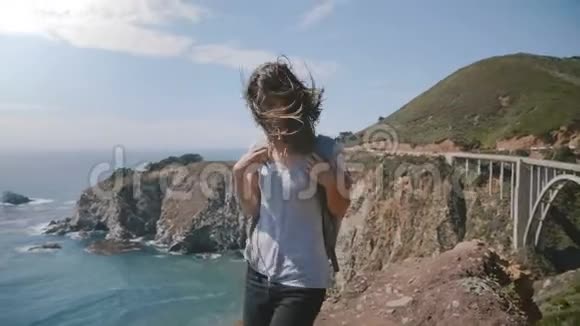 迷人的年轻女子与飞扬的头发和背包步行在令人惊叹的夏季海洋远景点在比克斯比桥大苏尔视频的预览图