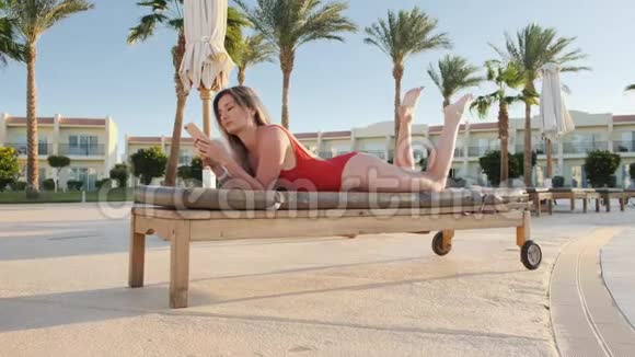 游泳池边的躺椅上躺着一位身穿红色泳装的美女一边在智能手机上浏览互联网视频的预览图