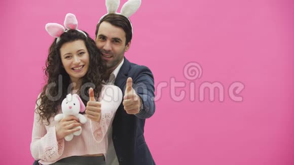 年轻夫妇站在粉红色的背景上在这个节目中一个手势课微笑着手里拿着一个柔软的玩具视频的预览图