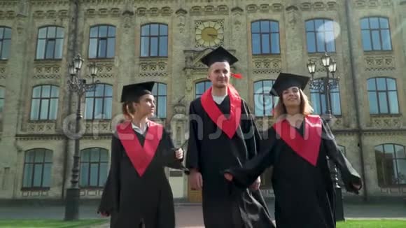 三个穿着黑色和红色壁炉的快乐学生在大学大楼前庆祝他们的毕业两个女孩视频的预览图