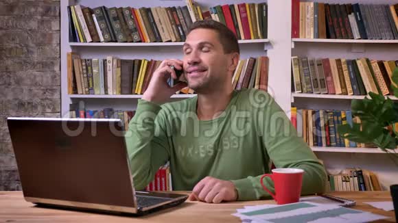 办公室工作人员坐在笔记本电脑前用温暖的面部表情在电话上交谈后面有书架视频的预览图