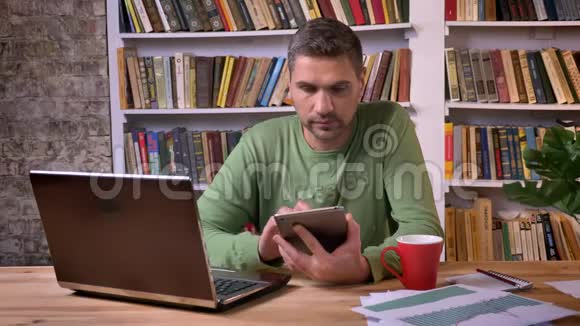 商人坐在笔记本电脑前喝着咖啡在平板电脑上滚动页面书架上的书架视频的预览图