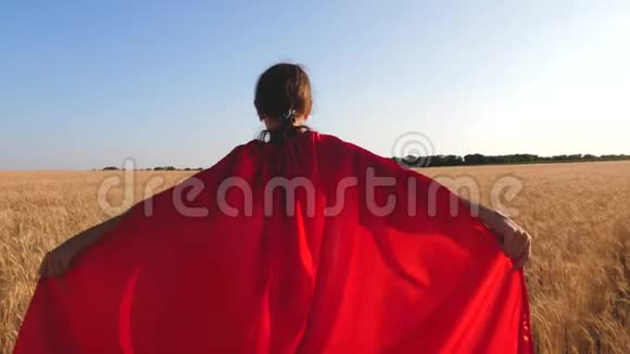 年轻女孩扮演超级英雄穿着红色雨衣的孩子穿过田野向蓝天跑去视频的预览图