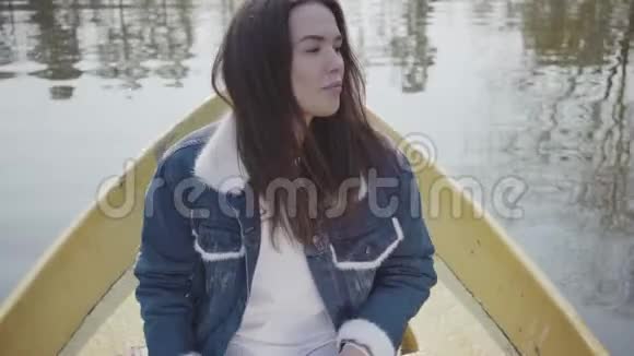 一位迷人的年轻女子穿着牛仔夹克漂浮在湖面或河流上的船上的肖像美丽的黑发视频的预览图