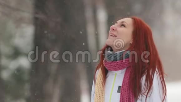 冬季公园站在人行道上的一个红头发的女人用嘴巴捕捉雪花慢动作视频的预览图