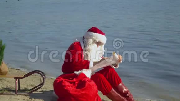 圣诞老人坐在湖边的沙滩上带着一个礼品袋在名单上写下一些东西视频的预览图