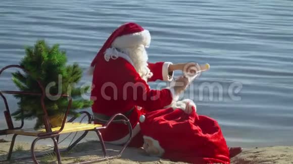 圣诞老人坐在湖边的沙滩上带着一个礼品袋在名单上写下一些东西视频的预览图
