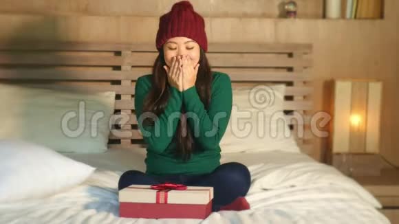 快乐美丽的亚洲女人戴着冬帽坐在床上拿着圣诞礼物兴奋地收到圣诞礼盒视频的预览图