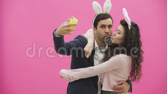 站在粉红色背景上的年轻嫩嫩的一对看着手机一边用手机自拍视频的预览图