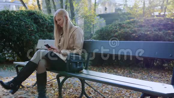 美丽的女孩穿着时尚的夹克在秋天公园的晴天在公园里使用智能手机视频的预览图