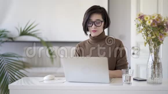 戴眼镜的亚洲女性在白色的家庭办公室里用笔记本电脑工作视频的预览图