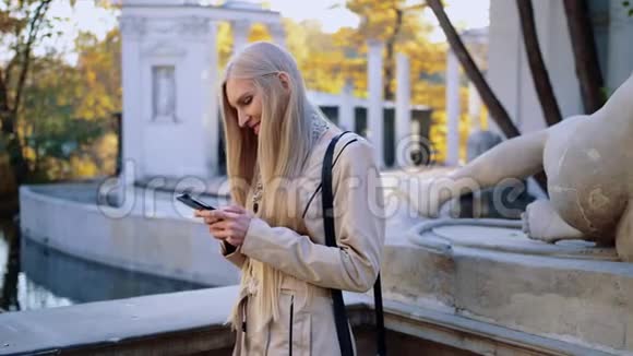秋公园里一位身穿米色外套的金发美女看着她的智能手机视频的预览图