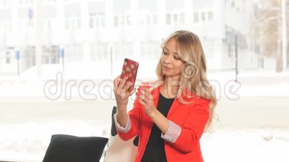 美丽的白种人年轻女子穿着红色外套在室内对着窗户自拍视频的预览图