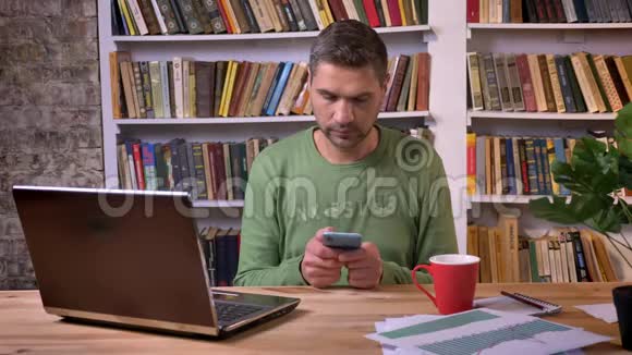 商人坐在笔记本电脑前在手机上浏览消息背景书架视频的预览图