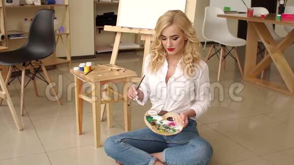 一个年轻的女孩有一个调色板和刷子坐在艺术工作室的地板上视频的预览图