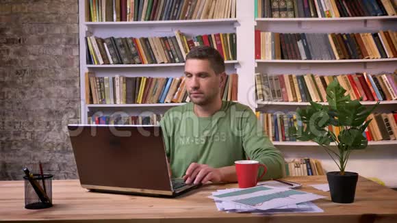 商人在笔记本电脑上工作喝咖啡并试图集中精力在办公室背景书架视频的预览图