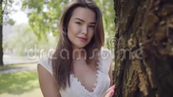 肖像可爱的年轻女孩一头长长的黑发穿着白色的夏装站在一棵树旁边视频的预览图