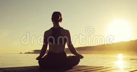 日落时分一位年轻女子坐在海边的码头上以莲花的姿势冥想视频的预览图