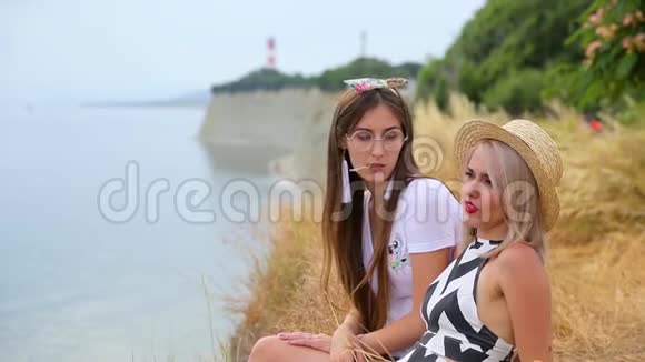 可爱的美丽苗条的年轻女子金发和黑发女孩坐在无边无际的蓝天下的海边悬崖上视频的预览图