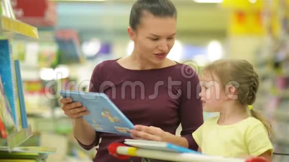 年轻的母亲和可爱的女儿在购物车选择儿童书籍在超市母亲节视频的预览图