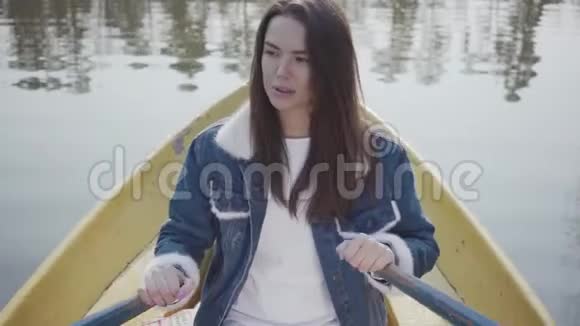 一个迷人的女孩穿着牛仔夹克漂浮在湖面或河流上的船上的肖像很漂亮的黑发女人视频的预览图