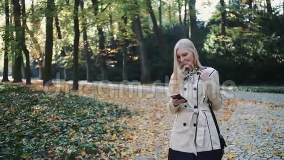 金碧辉煌的女子走在秋天的公园里看着智能手机和打字视频的预览图