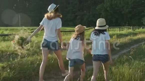 孩子们三个戴着帽子的女孩手牵着手沿着乡间小路跑回来视频的预览图