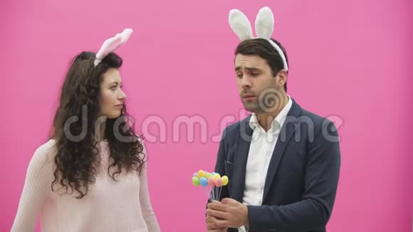 年轻夫妇站在粉红色的背景上在此期间他的妻子低下头手里拿着复活节彩蛋视频的预览图