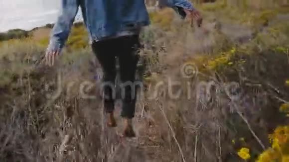 镜头跟随年轻快乐的女人在黄色的花朵和干燥的植物中漫步到大苏尔海海岸的神奇岩石上视频的预览图
