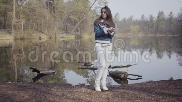 穿着白色裤子牛仔裤夹克和太阳镜站在河岸上的魅力女人女孩很冷她揉着她视频的预览图