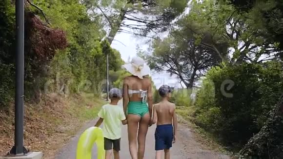 后景妈妈和两个儿子正在海边的公园里散步一个穿着泳衣戴着大帽子的年轻女人视频的预览图