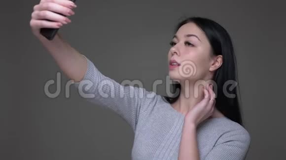 年轻漂亮的黑发白种人女性在手机上自拍的特写镜头背景是灰色的视频的预览图
