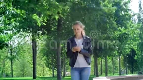 年轻漂亮的女孩正拿着智能手机在公园里散步读了留言很开心跳舞和弹跳视频的预览图