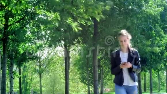 年轻漂亮的女孩正拿着智能手机在公园里散步读了留言很开心跳舞和弹跳视频的预览图