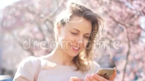 一位美丽的金发碧眼的春天肖像手里拿着一个电话背景是盛开的木兰树视频的预览图