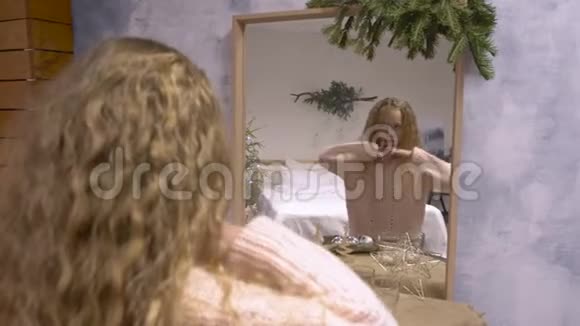 穿着粉色毛衣的年轻女孩在镜子前玩耍在圣诞树枝背景下头上长卷发视频的预览图