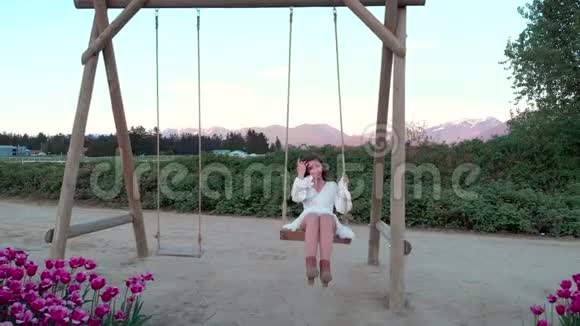 日落时分一位身穿白色夏装在郁金香花田附近荡秋千的年轻黑发女子视频的预览图