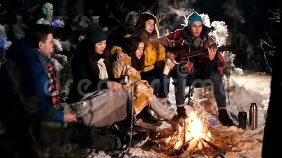 一群快乐的朋友坐在冬天的森林里吃着棉花糖一个弹吉他的年轻人温暖视频的预览图
