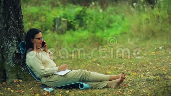 戴着眼镜的微笑着的女人光着脚坐在公园的一棵树下用铅笔在笔记本上写字然后继续说话视频的预览图
