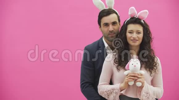 粉红色背景的年轻创意夫妇头上戴着黑黑的耳朵在这期间一个妻子抱着一只软玩具兔子一只视频的预览图